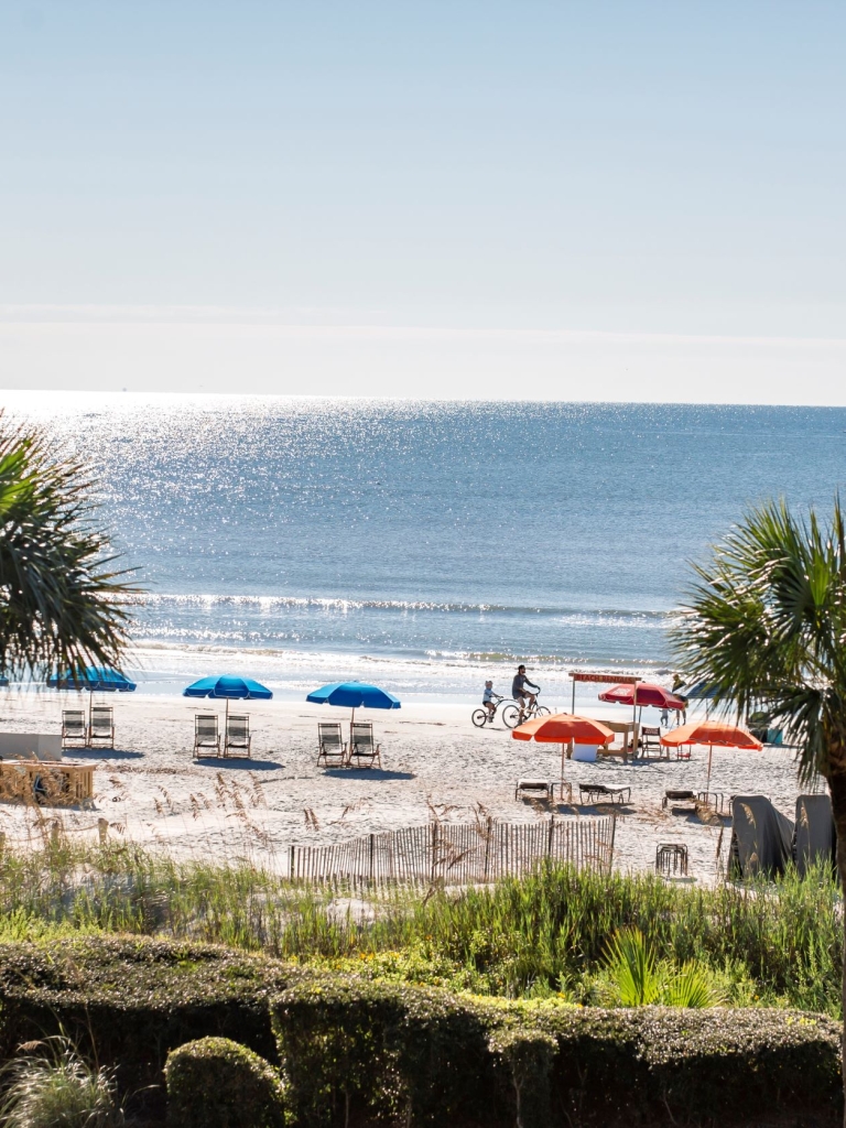 The view from the Sea Pines Beach Club of the beach with blue and orange umbrellas and a lifeguard stand and the ocean. 